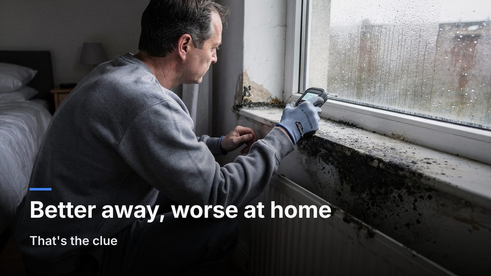 Person crouching at a damp bedroom window sill holding a small humidity meter near condensation on the glass